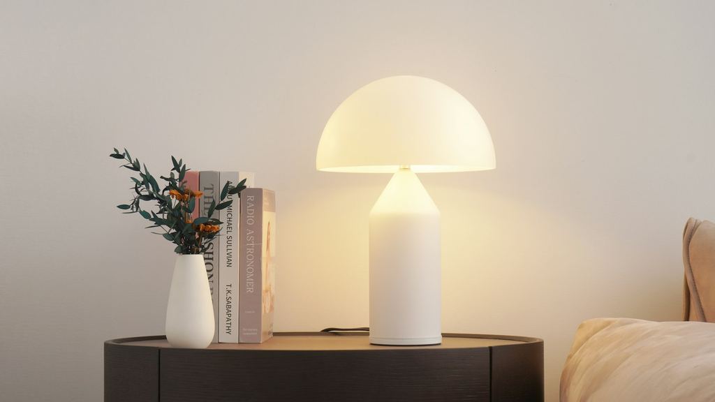 Tänd vit bordslampa med varmt sken på ett sidobord bredvid en vas och böcker